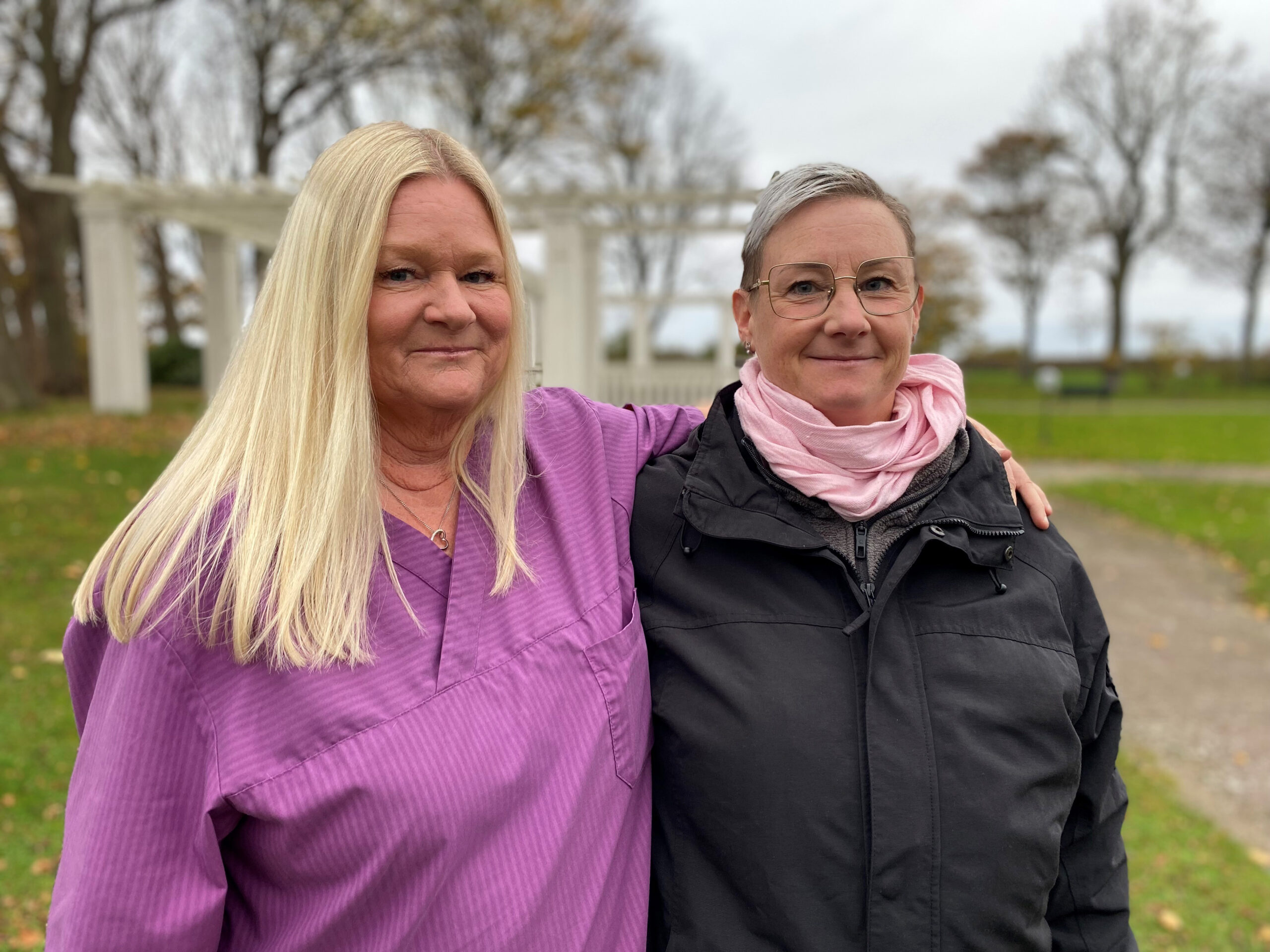 Två kvinnor står utomhus i en lummig grön miljö. Den ena kvinnan håller sin arm över den andra kvinnas axlar. Båda tittar in i kameran och ler. 