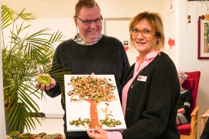 Två personer håller upp målad sten och tavla, tittar nöjt in i kameran.