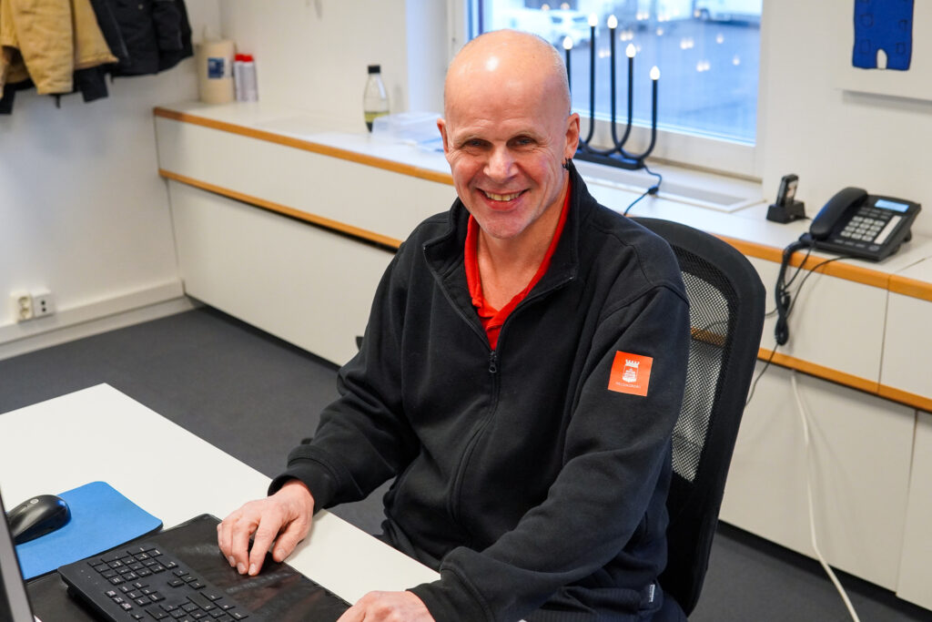 En man sitter vid ett skrivbord, tittar in i kameran och ler.