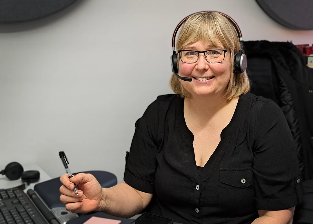 Kvinna med ljust halvlångt hår och glasögon som sitter vid ett skrivbord. Hon har headset på sig.