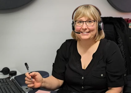 Kvinna med ljust halvlångt hår och glasögon som sitter vid ett skrivbord. Hon har headset på sig.