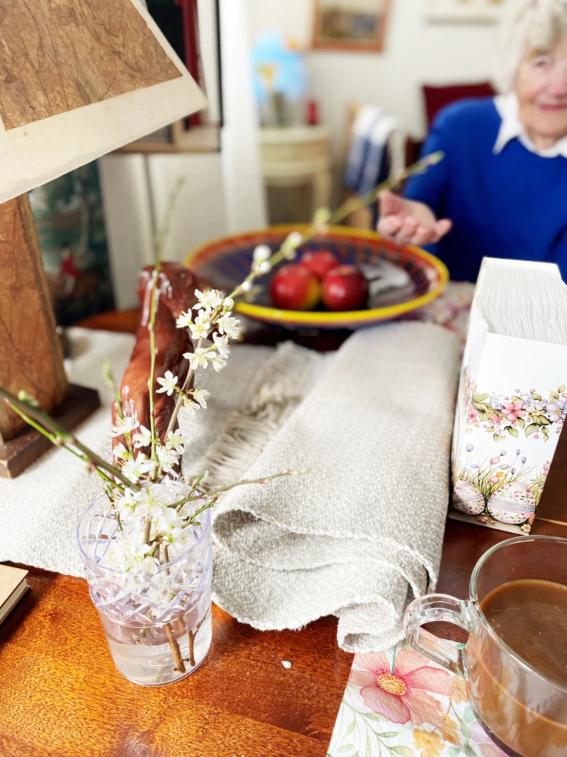 I förgrunden syns en kvist med blommor i en vas, i bakgrunden en äldre dam som pekar på vasen