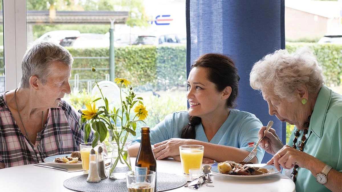 Ung kvinna sitter på huk intill två äldre damer som äter lunch