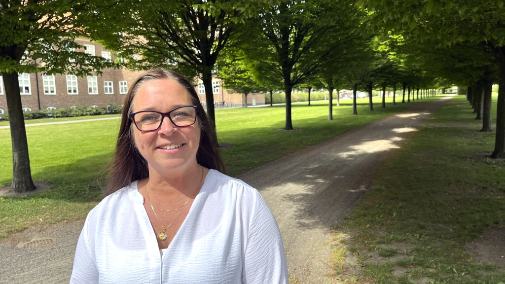 Mörkhårig kvinna i glasögon står framför en grönskande allé och tittar in i kameran