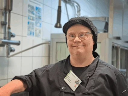 En kvinna står i ett diskrum. hon har svarta kläder, svart keps och glasögon.