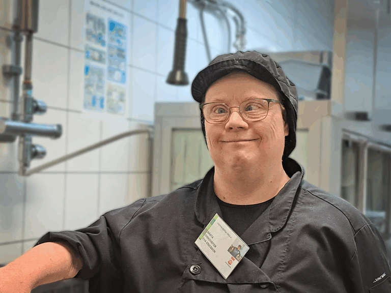 En kvinna står i ett diskrum. hon har svarta kläder, svart keps och glasögon.