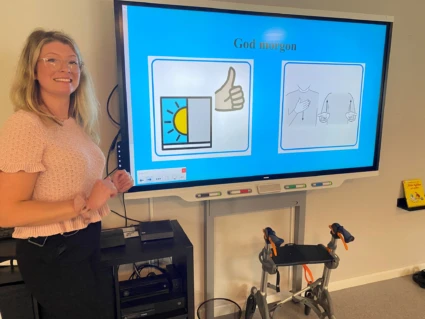 Person står bredvid skärm, ler och tittar in i kameran.