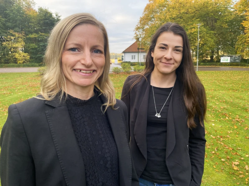 Två kvinnor står utomhus, klädda i svarta kläder, tittar in i kameran och ler. 