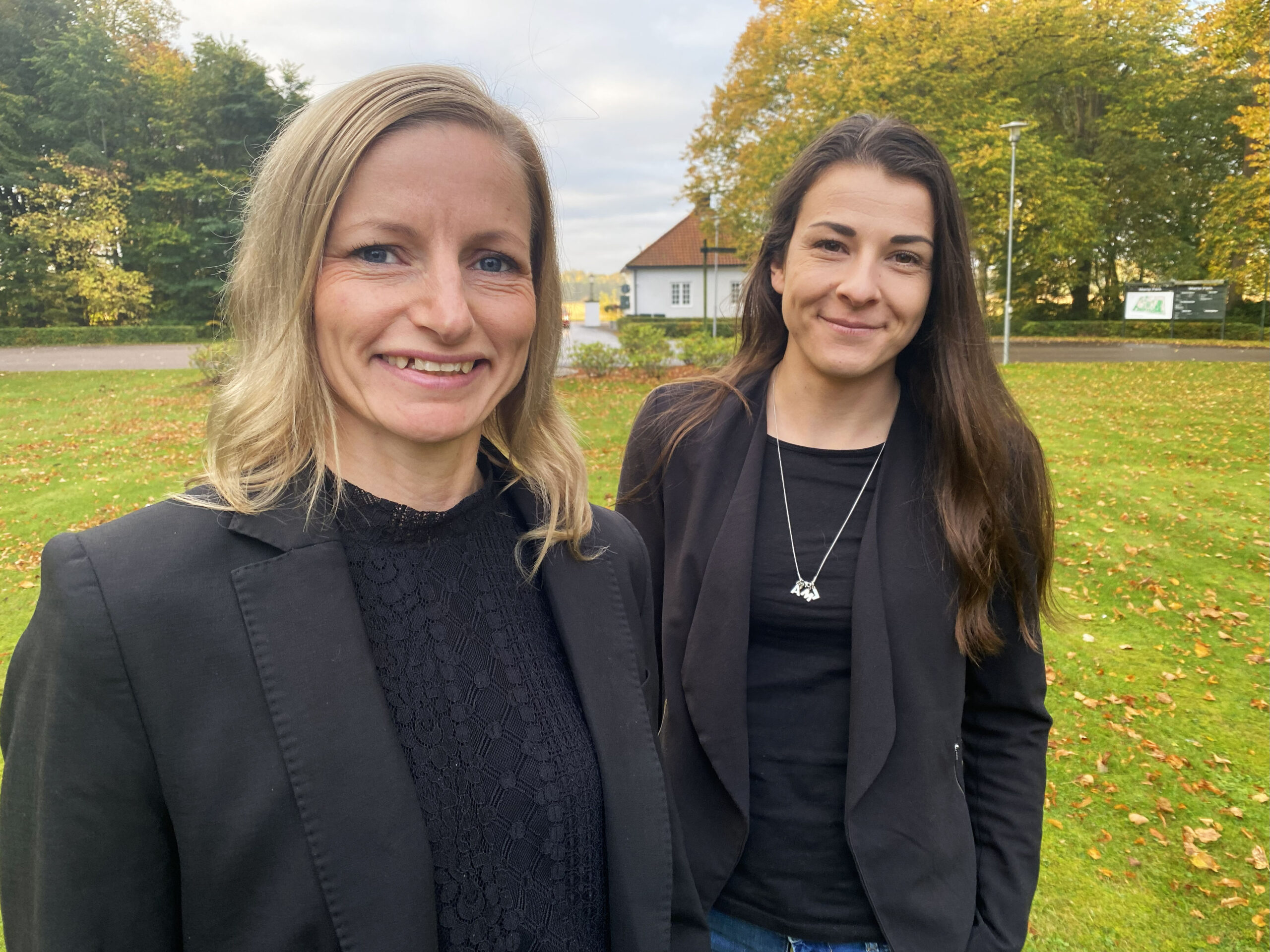 Två kvinnor står utomhus, klädda i svarta kläder, tittar in i kameran och ler.