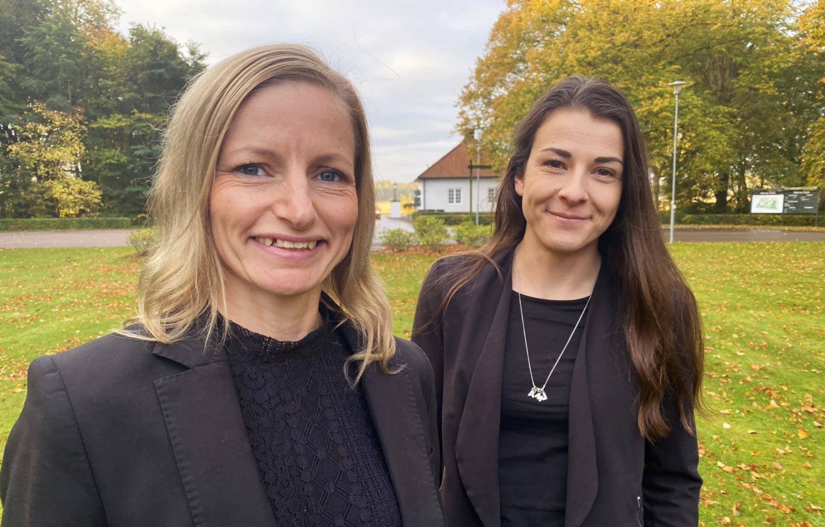 Två kvinnor står utomhus, de bär svarta kläder, tittar in i kameran och ler.