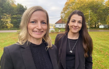 Två kvinnor står utomhus, de bär svarta kläder, tittar in i kameran och ler.