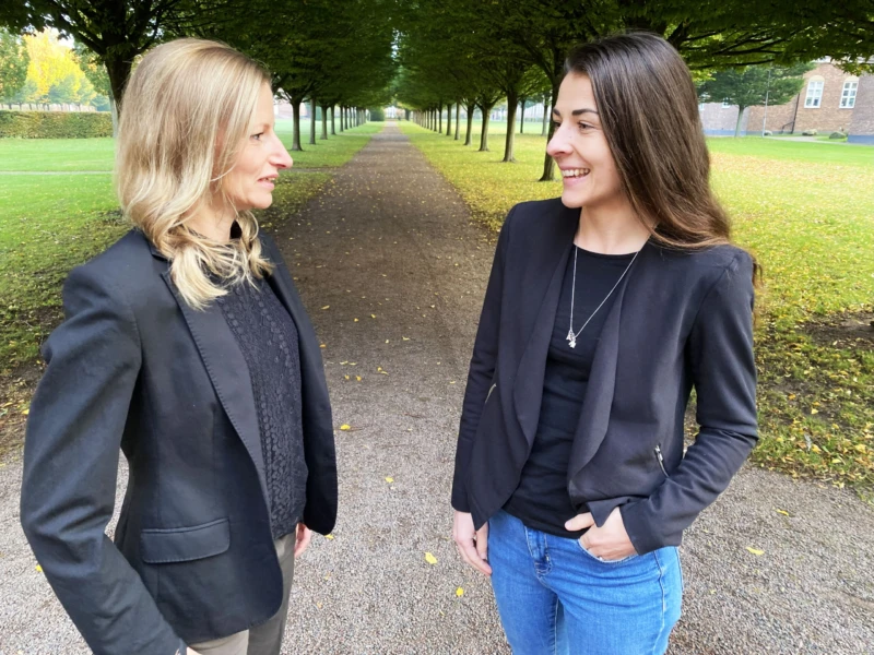 Två kvinnor, klädda i svart, står utomhus, tittar på varandra och ler. 