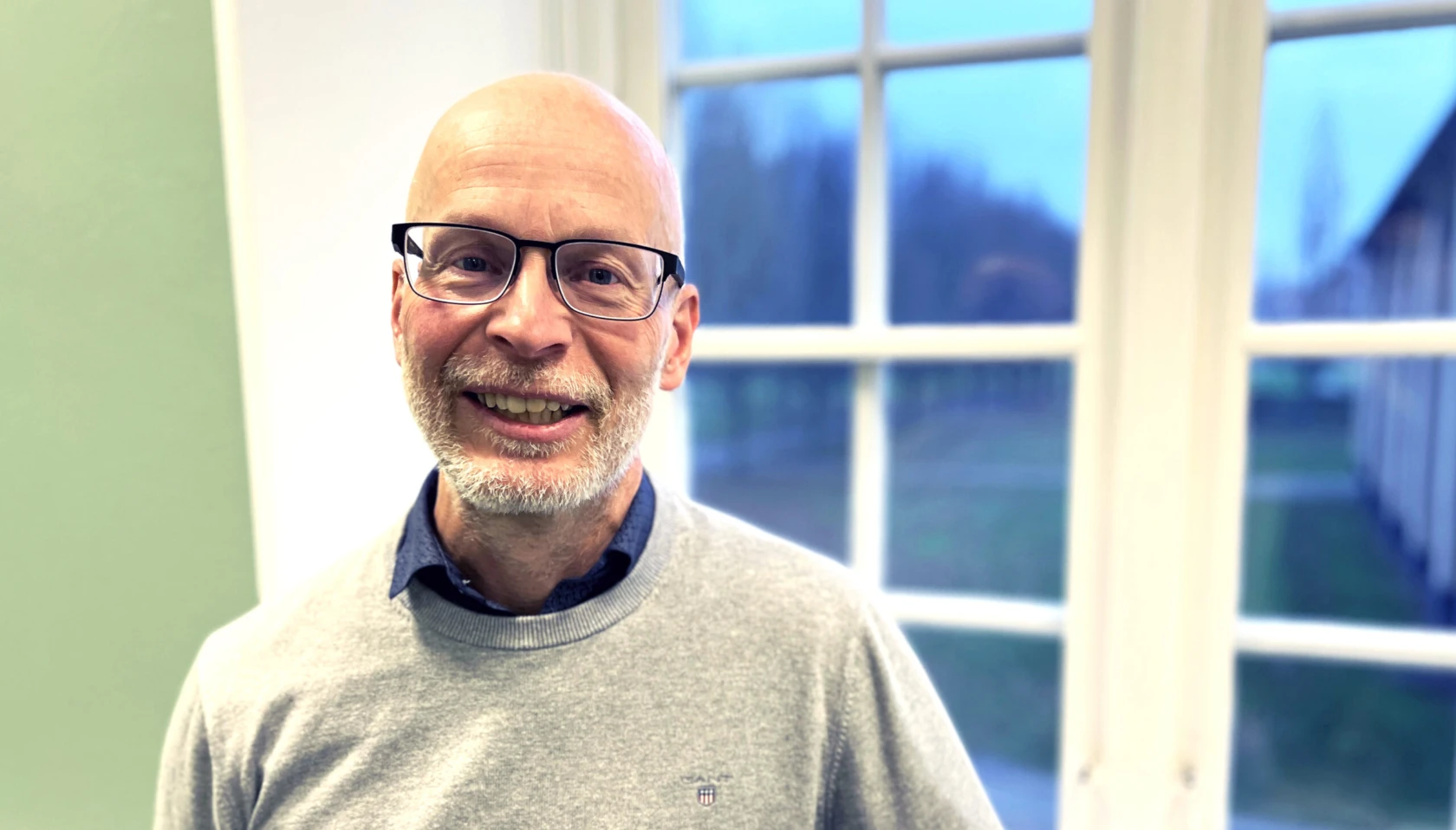 Medelålders man med skägg står framför ett fönster och tittar in i kameran.