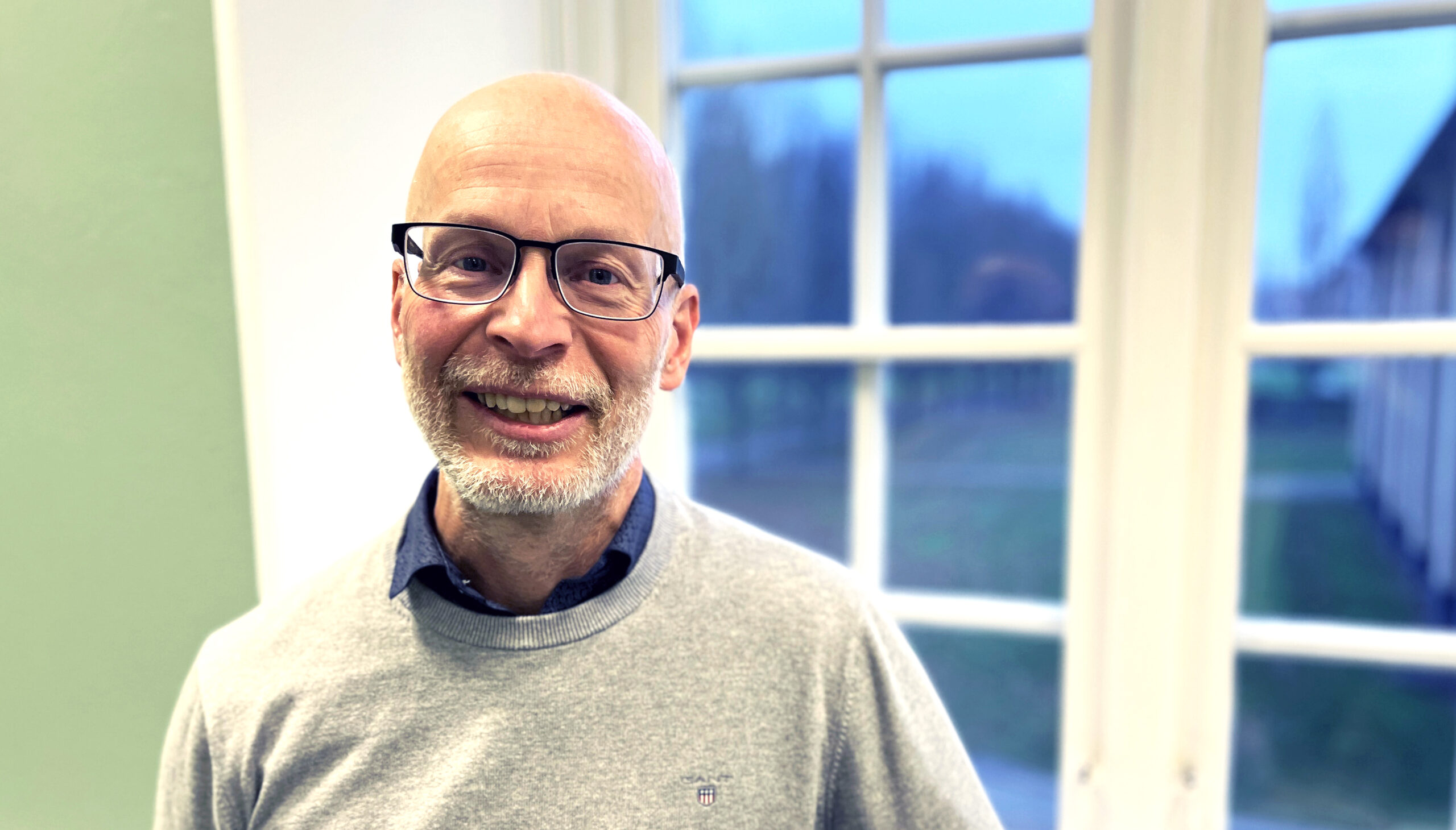 Medelålders man med skägg står framför ett fönster och tittar in i kameran.