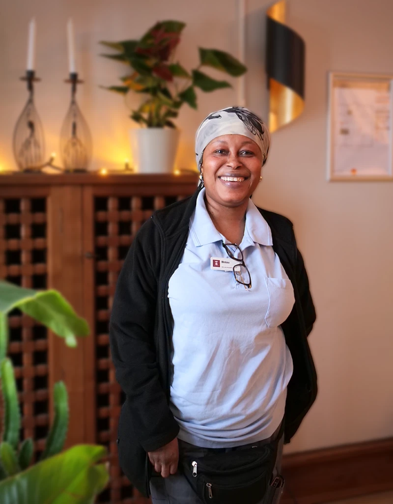 En kvinna står inomhus och ler mot kameran. Hon bär ljusblå arbetskläder, svart kofta och en mönstrad huvudduk. I bakgrunden syns en byrå med ljusstakar och en grön växt.