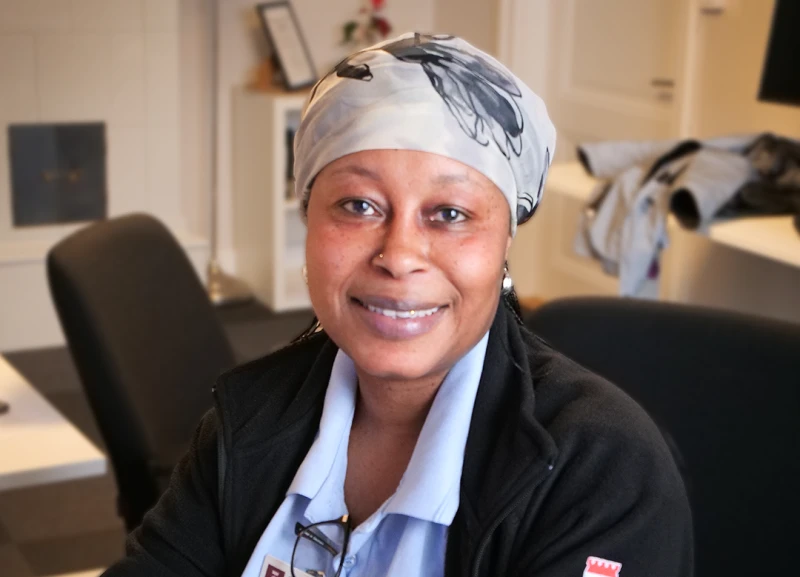 Kvinna som sitter vid ett bord och ler mot kameran. Hon bär ljusblå arbetskläder, svart kofta och en mönstrad huvudduk.