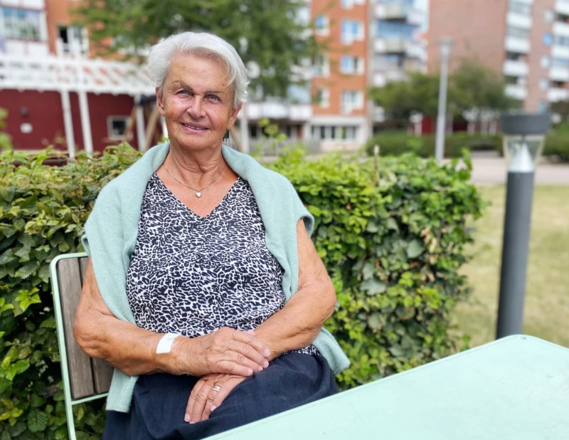 Äldre kvinna sitter vid ett bord utomhus. Hon tittar in i kameran och ler. 