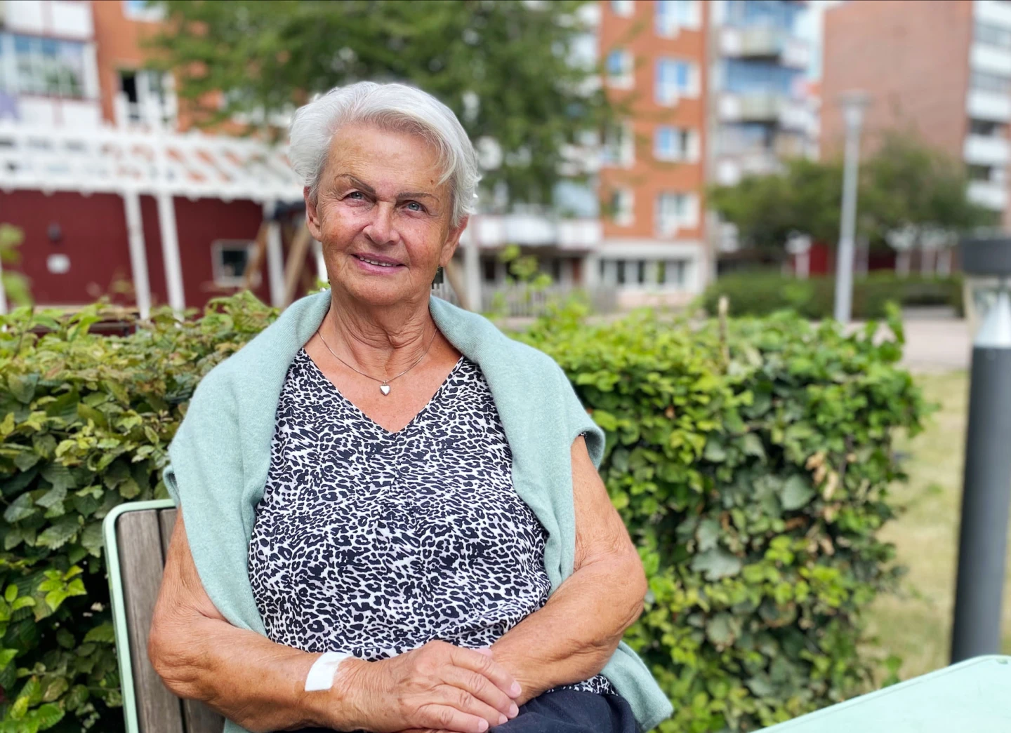 Äldre kvinna sitter vid ett bord utomhus. Hon tittar in i kameran och ler.
