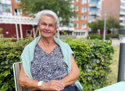 Äldre kvinna sitter vid ett bord utomhus. Hon tittar in i kameran och ler.