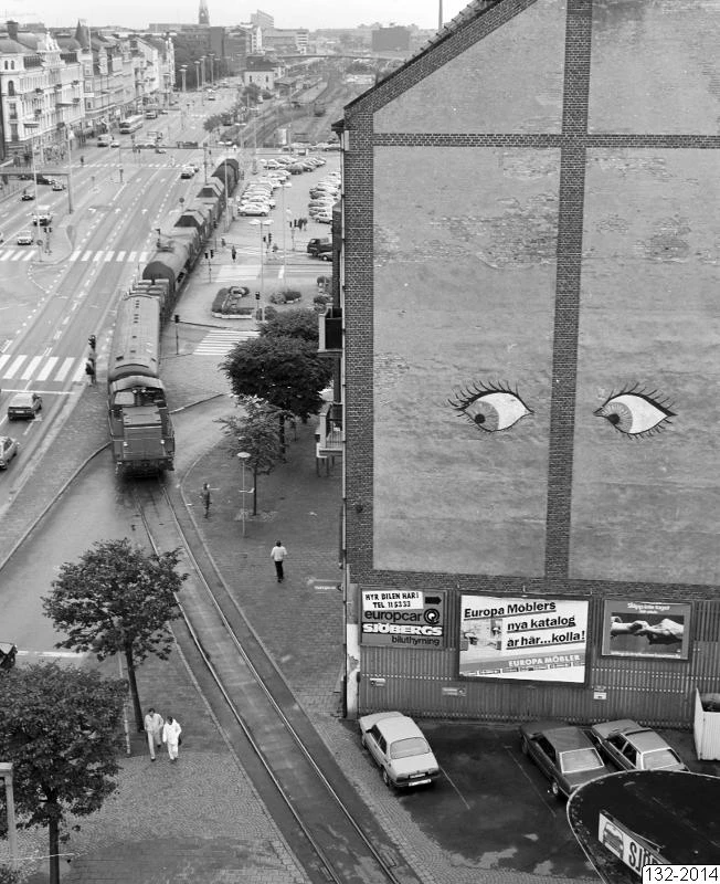 En byggnad “ser på” när ett godståg rullar genom centrala Helsingborg över Järnvägsgatan på väg till Norra Hamnen. I bakgrunden syns den gamla Centralstationen och Södra Hamnens bangård. Källa: https://museum.helsingborg.se/web/object/760047