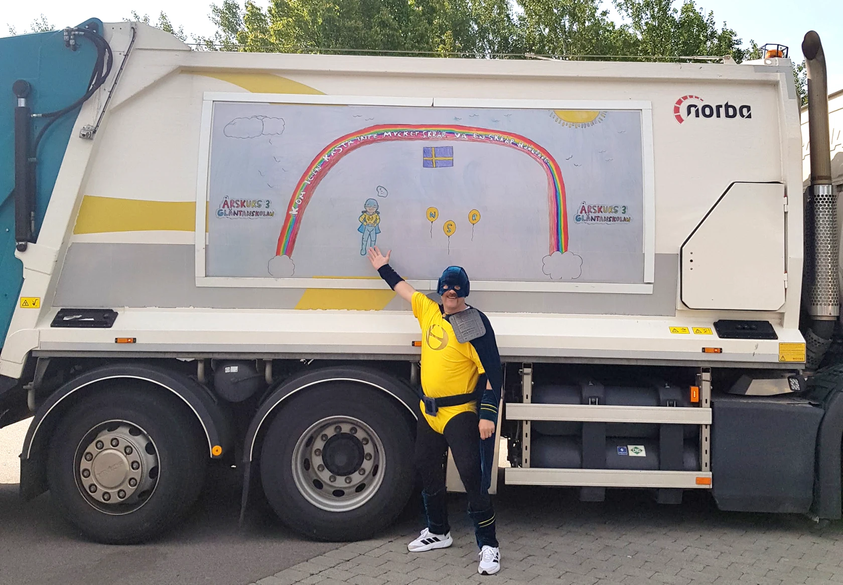 Sophjälte med ljusblå mask och gul dräkte visar en regnbågsteckning på en sopbil.