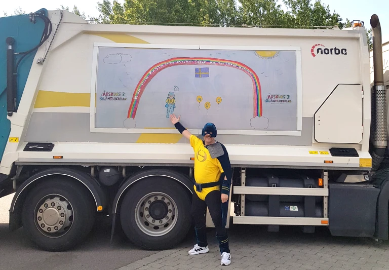 Sophjälte med ljusblå mask och gul dräkte visar en regnbågsteckning på en sopbil.
