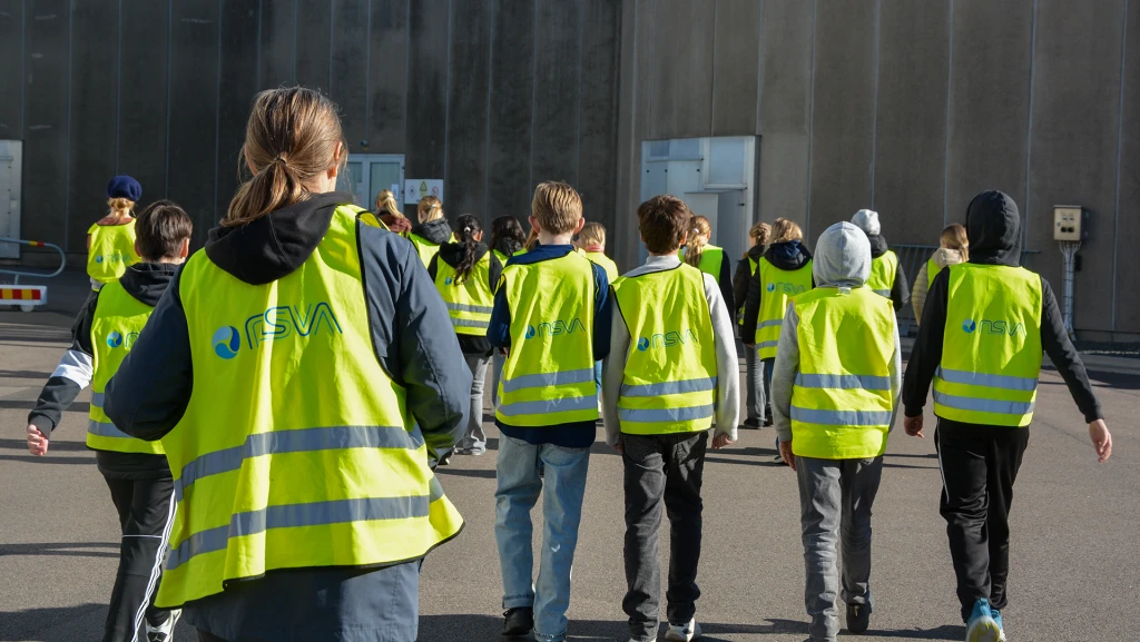 En grupp elever och en lärare med gula besöksvästar går mot en grå betongbyggnad.