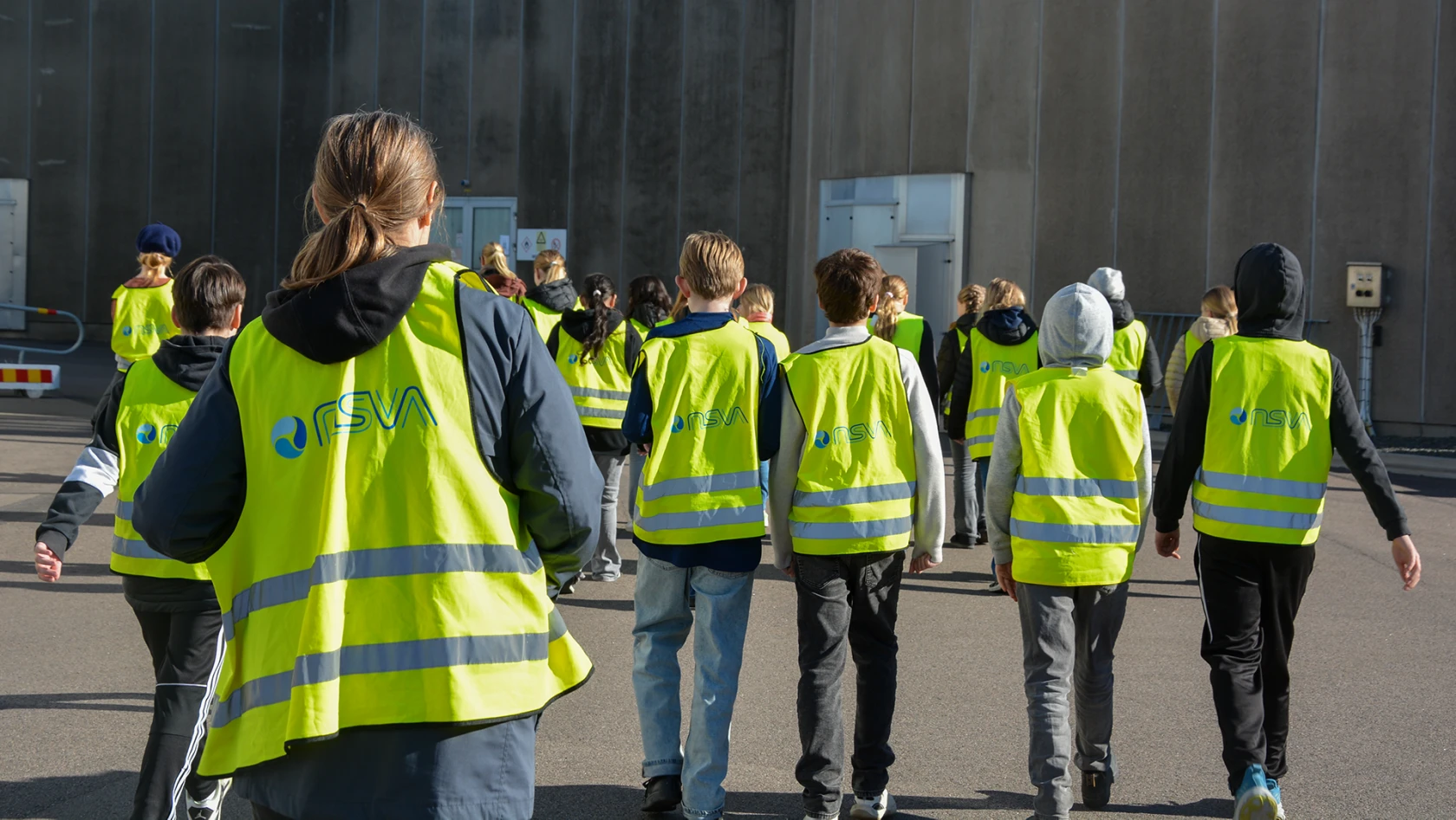 En grupp elever och en lärare med gula besöksvästar går mot en grå betongbyggnad.