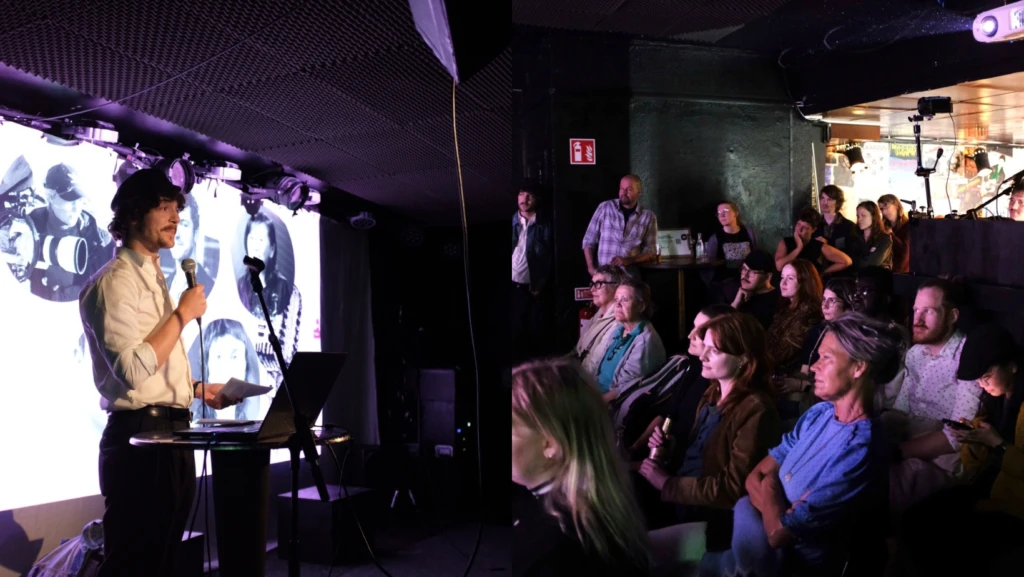 En talare står på scenen med mikrofon och håller ett papper, framför en stor skärm som visar bilder på filmskapare. Publiken, som sitter och står framför scenen i ett mörkt belyst rum, lyssnar uppmärksamt. En projektor och ljudutrustning syns i lokalen.