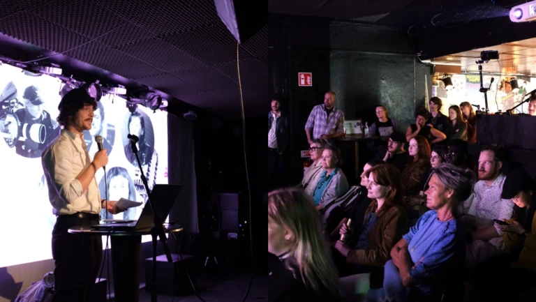 En talare står på scenen med mikrofon och håller ett papper, framför en stor skärm som visar bilder på filmskapare. Publiken, som sitter och står framför scenen i ett mörkt belyst rum, lyssnar uppmärksamt. En projektor och ljudutrustning syns i lokalen.