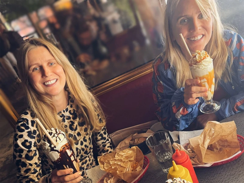 Amanda och Adina sitter vid ett bord på en restaurang och dricker milkshakes skrattandes.