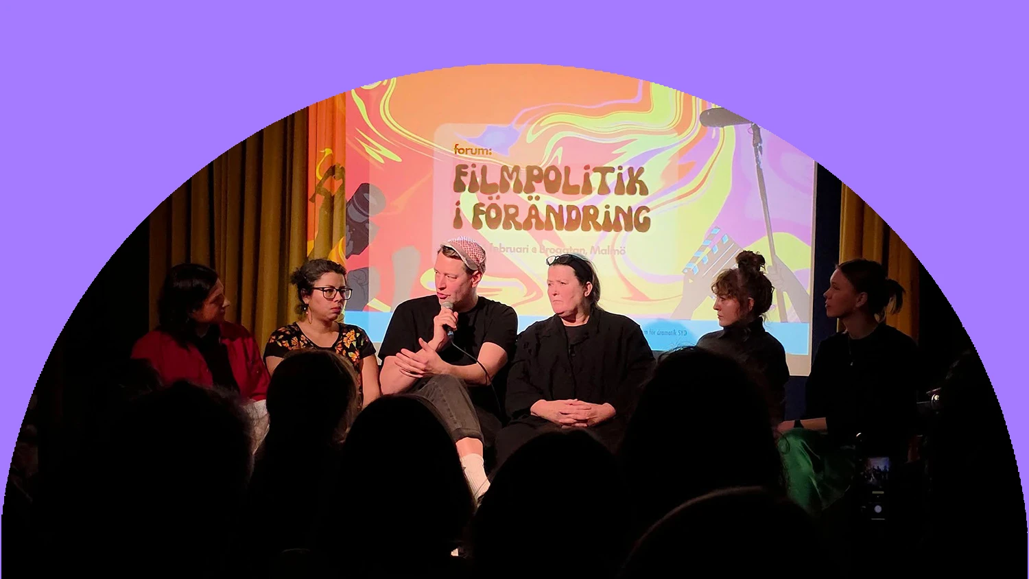 Personer sitter bredvid varandra på en scen. En talar i en mikrofon medan de andra tittar på.