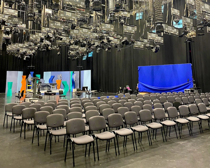 Stor sal med många stolar på rad. I bakgrunden syns scenografier och i taket hänger massor av studiolampor.